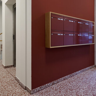 Ein heller, moderner Flur im Eingangsbereich eines Gebäudes. An einer rotbraunen Wand hängt eine hölzerne Briefkastenanlage mit rotbraunen Klappen. Der Boden ist aus rötlich-weiß gesprenkeltem Terrazzo. Links führt der Gang zu einem Aufzug und einer Treppe mit Holzhandlauf.