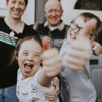 Eine fröhliche vierköpfige Familie vor einer weißen Haustür. Im Vordergrund zeigen ein Mädchen und ein Junge mit Brille lachend ihre Daumen nach oben. Hinter ihnen stehen die lächelnden Eltern, die ebenfalls den Daumen heben.