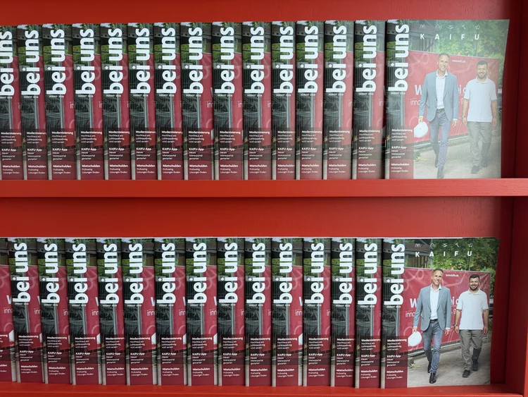 Zwei Regalböden eines knallroten Regals sind dicht an dicht mit Ausgaben des Magazins „bei uns“ gefüllt. In beiden Reihen stehen die Hefte seitlich gestapelt, sodass man vor allem den Schriftzug „bei uns“ auf den Magazinrändern sieht. Auf der jeweils rechten Seite liegt das Cover flach auf: Es zeigt zwei Männer vor einem Gebäude.