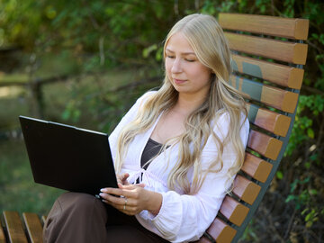 Eine junge blonde Frau sitzt draußen auf einer Holzliege im Grünen und arbeitet konzentriert an einem Laptop.