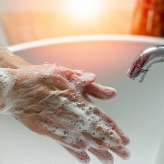 Nahaufnahme von zwei Händen, die sich gründlich unter weißem Seifenschaum reiben, aufgenommen über einem weißen Waschbecken neben einem verchromten Wasserhahn.
