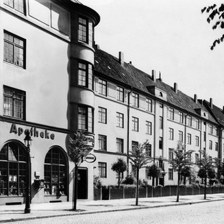 Historische Schwarz-Weiß-Aufnahme einer gepflasterten Wohnstraße. Links befindet sich ein mehrstöckiges Gebäude mit der großen Aufschrift „Apotheke“ und Rundbogenfenstern im Erdgeschoss. Entlang des Gehwegs erstreckt sich eine lange Häuserzeile, vor der eine Reihe junger Bäume gepflanzt ist.