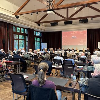 Blick von hinten in einen gut gefüllten Tagungssaal mit Holzgebälk an der Decke während einer Versammlung. Die Teilnehmer sitzen an Tischen mit Blick auf das Podium an der Stirnseite. Dort sitzt ein Podiumsteam vor einer großen Leinwand, auf der eine rote Präsentationsfolie mit der Aufschrift „TOP 11 Verschiedenes“ projiziert wird. Links geben große Fensterfronten den Blick ins Grüne frei, rechts stehen Roll-up-Banner der Genossenschaft.