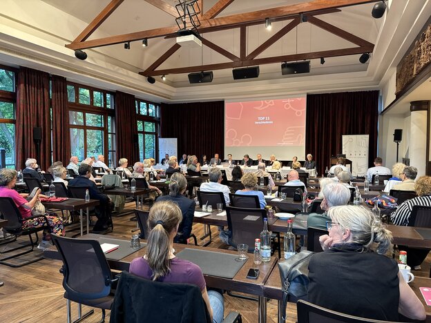 Blick von hinten in einen gut gefüllten Tagungssaal mit Holzgebälk an der Decke während einer Vertreterversammlung. Die Teilnehmer sitzen an Tischen mit Blick auf das Podium an der Stirnseite. Dort sitzt ein Podiumsteam vor einer großen Leinwand, auf der eine rote Präsentationsfolie mit der Aufschrift „TOP 11 Verschiedenes“ projiziert wird. Links geben große Fensterfronten den Blick ins Grüne frei, rechts stehen Roll-up-Banner der Genossenschaft.