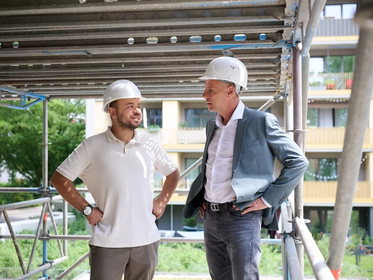 Zwei Männer stehen auf der Plattform eines Metallgerüsts vor einem modernen Mehrfamilienhaus. Beide tragen weiße Schutzhelme. Der linke Mann trägt ein helles Poloshirt und hat die Hände in die Hüften gestützt. Der rechte Mann trägt ein weißes Hemd, ein graues Sakko sowie eine dunkle Hose und schaut den anderen Mann an. Im Hintergrund ist die Fassade eines Wohnhauses mit Balkonen zu sehen.