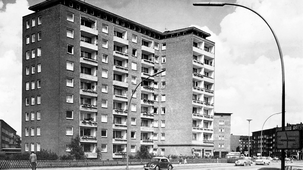 Eine Schwarz-Weiß-Postkarte, die ein modernes Wohnhochhaus in den 1960er Jahren zeigt. Es handelt sich um ein l-förmiges Gebäude mit Balkonen, das an einer mehrspurigen Straße liegt.