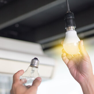 Eine Hand schraubt eine leuchtende LED-Lampe in eine Fassung an der Decke, während die andere Hand eine alte, durchsichtige Glühbirne hält.