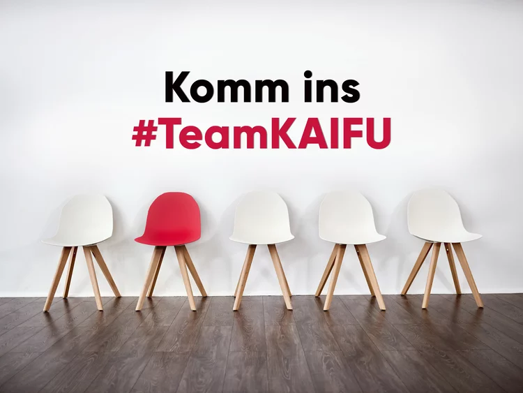 Eine Reihe von fünf Stühlen vor einer weißen Wand mit der Aufschrift „KOMM INS #TEAMKAIFU“. Vier der Stühle sind weiß, während einer in kräftigem Rot hervorsticht.