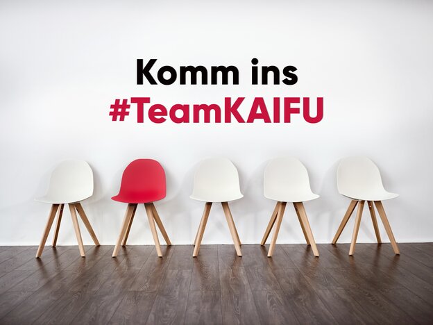 Une rangée de cinq chaises devant un mur blanc portant l'inscription « KOMM INS #TEAMKAIFU ». Quatre de ces chaises sont blanches, tandis qu'une autre se détache par sa couleur rouge vif.