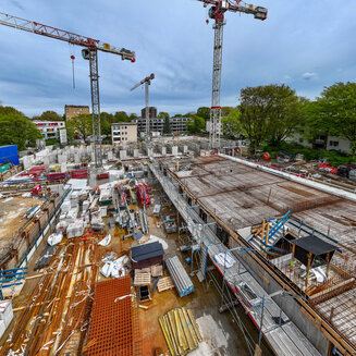Vogelperspektive auf eine weitläufige Großbaustelle für einen Gebäudekomplex. Im Vordergrund liegt eine große, frisch verlegte Deckenfläche aus Bewehrungsmatten (Bewehrungsstahl). Auf dem Areal verteilt stehen mehrere große Turmdrehkräne. Am Boden lagern Baumaterialien wie rostige Stahlgitter, Holzlatten, Gerüstteile und blaue Baucontainer. Im Hintergrund sind umliegende grüne Bäume und angrenzende Wohngebäude unter einem leicht bewölkten Himmel zu sehen.