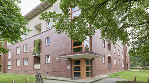 Eckansicht eines rotbraunen Klinker-Wohngebäudes mit überdachtem Haupteingang und großen Fensterfronten im Treppenhaus.