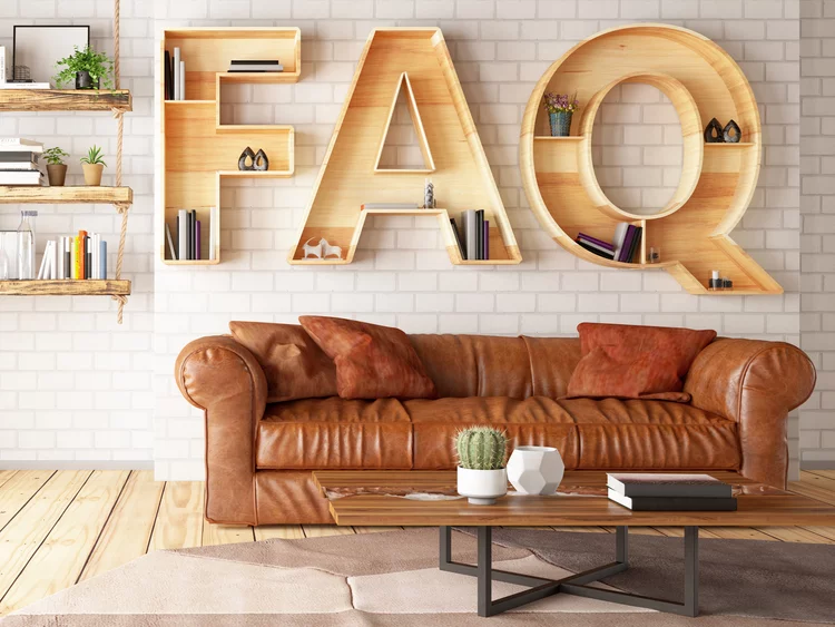 Ein modernes Wohnzimmer im Loft-Stil. An einer weißen Backsteinwand hängen Wandregale in Form der großen Buchstaben „FAQ“, die mit Büchern und Deko gefüllt sind. Darunter steht ein braunes Ledersofa. Links stehen ein Hängeregal und ein schwarzes Fahrrad an einem großen Fenster.