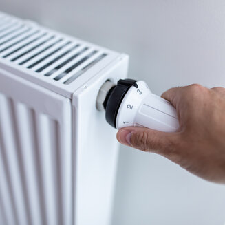 Nahaufnahme eines weißen Flachheizkörpers an einer hellen Wand. Eine Hand fasst von rechts nach dem Thermostatkopf mit den Zahlen 1 bis 3, um die Heizung einzustellen. Der Hintergrund ist leicht unscharf gehalten.