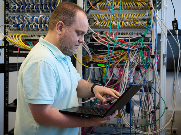 Ein Mann mit kurzen Haaren, Dreitagebart und einem hellblauen Poloshirt steht im Profil vor einem Server- und Netzwerk-Rack. Er hält einen geöffneten, schwarzen Laptop in den Händen und bedient aufmerksam das Touchpad oder die Tastatur. Der Serverraum im Hintergrund ist gefüllt mit verkabelten Netzwerk-Switches und Patchpanels, aus denen zahlreiche bunte Netzwerkkabel (gelb, grün, blau, rosa) verlaufen.