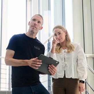 Ein Mann mit Tablet und eine Frau in einer hellen Bluse stehen auf einer Treppe in einem modernen Gebäude und blicken nach oben, während die Frau in eine Richtung zeigt.
