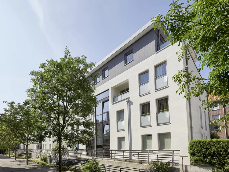 Außenaufnahme eines modernen Mehrfamilienhauses unter blauem, leicht bewölktem Himmel. Die hell verputzte Fassade verfügt über ein dunkelgrau abgesetztes Staffelgeschoss, große Fenster mit silbernen Geländern und ein zentrales Treppenhaus mit durchgehender Glasfront. Im Vordergrund führt eine Pflasterstraße an mehreren Laubbäumen und gepflegten Grünflächen vorbei. Ein Metallgeländer trennt den Gehweg von den Keller- und Eingangsbereichen der Anlage.