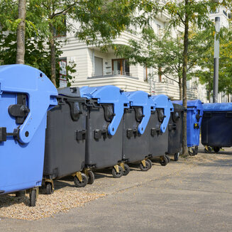 Mehrere große Abfallcontainer für Altpapier stehen in einer Reihe im Außenbereich. Die Tonnen haben dunkelgraue bis blaue Korpusse und durchgehend blaue Runddeckel. Im Hintergrund befinden sich Bäume und ein helles, mehrstöckiges Wohnhaus.
