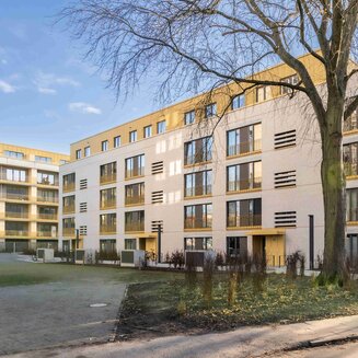 Außenaufnahme eines mehrgeschossigen, neugebauten Wohnhauses bei sonnigem Wetter. Die Fassade besteht aus hellbeigefarbenen Klinkersteinen, kombiniert mit einem obersten Geschoss und Balkongittern in einem warmen Goldgelb-Ton. Vor dem Gebäude steht ein großer, laubloser Baum. Im Vordergrund sind eine Pflasterfläche, kleine Sträucher sowie ein Gehweg zu sehen. Links im Hintergrund schließt sich ein weiteres modernes Wohngebäude an.