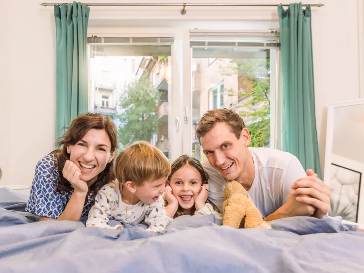 Eine vierköpfige Familie liegt glücklich lächelnd nebeneinander auf einem Bett im hellen Schlafzimmer vor einem Fenster mit grünen Vorhängen.