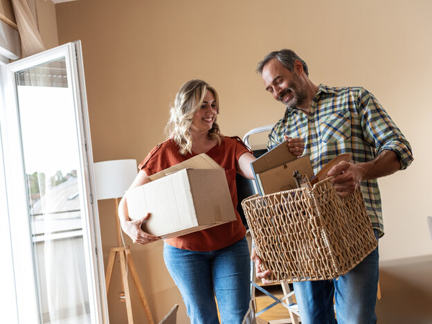 A man and a woman are standing in a living room, smiling, whilst moving house. The woman is carrying a moving box under her arm, whilst the man is holding a wicker basket, from which they are unpacking decorative items together.
