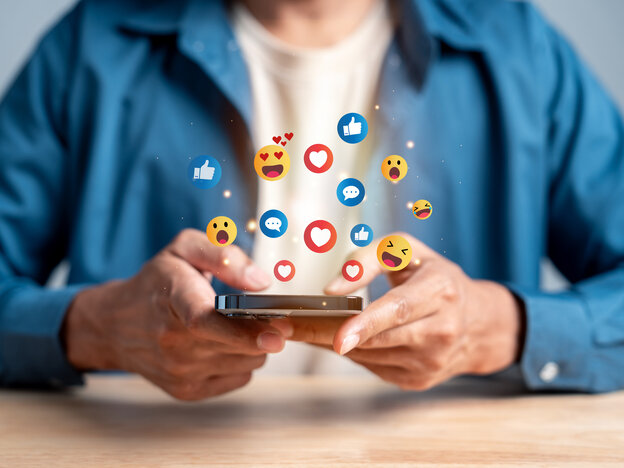 Eine Person hält ein Smartphone, über dem zahlreiche bunte Social-Media-Icons und Emojis schweben.