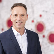Porträt von Stefan Henze im dunkelblauen Anzug und weißen Hemd vor einer hellen Wand mit roten Kreisen.