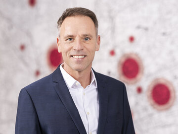 Porträt von Stefan Henze, technischem Vorstand der KAIFU-NORDLAND. Er trägt einen dunkelblauen Anzug mit weißem Hemd, lächelt und blickt direkt in die Kamera. Im Hintergrund ist eine helle Wand mit unscharfen roten Kreisen zu sehen.