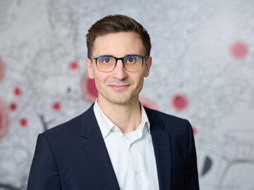 Ein junger Mann mit kurzen braunen Haaren, Eric Yeung-Schüürmann, blickt lächelnd in die Kamera. Er trägt eine Brille mit dunklem Rahmen, ein weißes Hemd und ein dunkelblaues Sakko. Im unscharfen Hintergrund ist ein schematischer Stadtplan in Grau mit mehreren roten Markierungspunkten zu sehen.