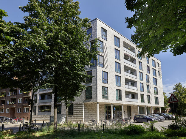Ein modernes, fünfstöckiges Wohnhaus aus grauem Klinker steht unter einem blauen Himmel, eingerahmt von großen, grünen Bäumen. Das Gebäude hat zahlreiche Balkone und große Fenster. Im Erdgeschoss befinden sich Säulen und eine Glasfront. Davor parken Autos, Motorräder und Fahrräder. Links ist ein älteres Backsteingebäude zu sehen.