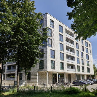Ansicht eines modernen, fünfstöckigen Wohnhauses mit heller Klinkerfassade und Balkonen in einer von Bäumen gesäumten Straße in Hamburg. Davor parken Autos und Fahrräder unter klarem blauen Himmel.