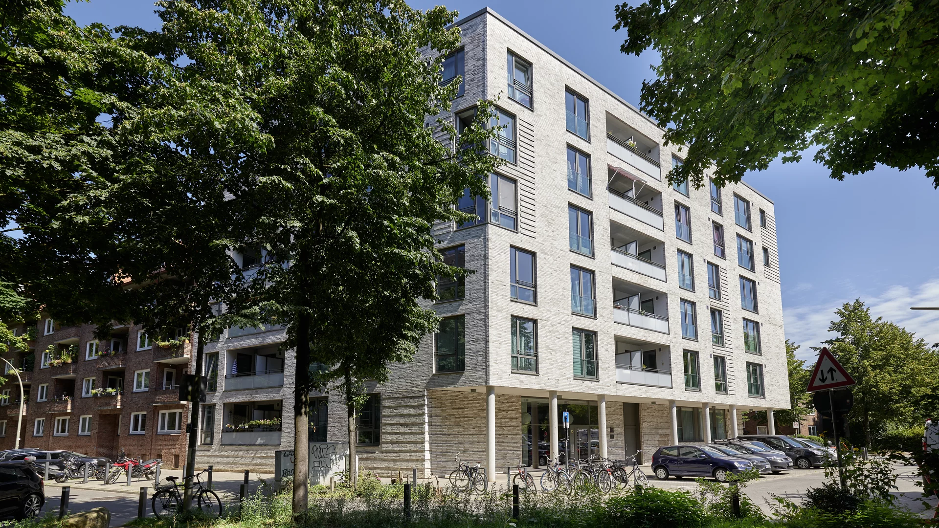 Eine Außenaufnahme eines modernen, fünfstöckigen Wohngebäudes in Hamburg an einem sonnigen Tag. Die Fassade ist mit hellen, horizontal strukturierten Riemchen verkleidet. Die oberen Stockwerke haben große Fenster und zurückgesetzte Balkone. Das Erdgeschoss steht auf schlanken Säulen und hat eine gläserne Eingangshalle. Mehrere große, grüne Bäume im Vordergrund beschatten den linken Teil des Gebäudes und den Gehweg. Am Straßenrand parken mehrere Autos (darunter ein blauer Kleinwagen) und viele Fahrräder sind an Fahrradständern angeschlossen. Rechts ist ein rotes Dreieck-Verkehrsschild (Vorfahrt gewähren) zu sehen. Im Hintergrund links ist ein älteres Backsteingebäude sichtbar.