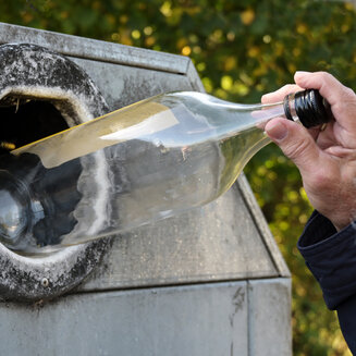 Nahaufnahme einer Hand, die eine transparente Glasflasche mit schwarzem Drehverschluss in die kreisrunde Einwurföffnung eines grauen Altglascontainers wirft. Im Hintergrund sind grüne Blätter zu erkennen.