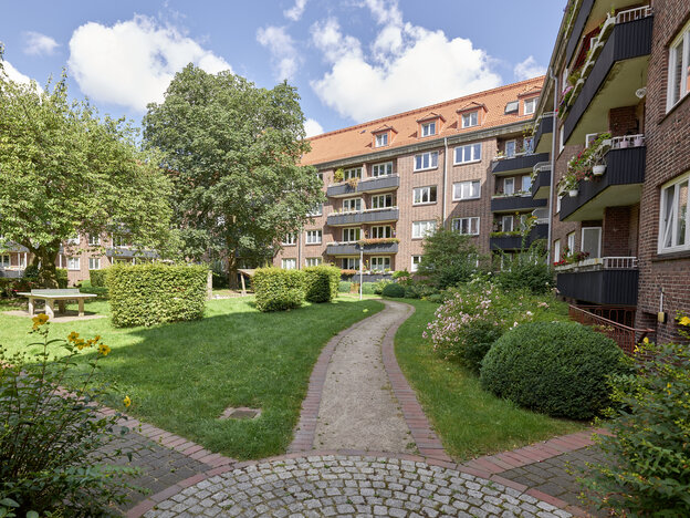 Sonnige Innenhof-Gartenanlage umgeben von mehrgeschossigen Rotklinker-Wohngebäuden mit Balkonen. Im Vordergrund verläuft ein gepflasterter Weg, umgeben von gepflegtem Rasen, Büschen, Blumen und Bäumen. Im Hintergrund steht eine Tischtennisplatte im Schatten eines großen Baumes.