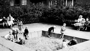 Eine historische Schwarz-Weiß-Fotografie eines belebten Kinderspielplatzes in einer Wohnanlage aus den 1960er Jahren. Mehrere Kinder spielen in einem großen, von Betonplatten eingefassten Sandkasten.