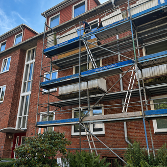 An einem roten Backsteinwohnhaus in der Neumünsterschen Straße steht ein mehrstöckiges Baugerüst. Ein Handwerker arbeitet auf der oberen Gerüstebene an den Balkonen, deren Verkleidungen teilweise entfernt wurden. Leitern verbinden die einzelnen Ebenen. Vor dem Gebäude wachsen Sträucher und Farne.