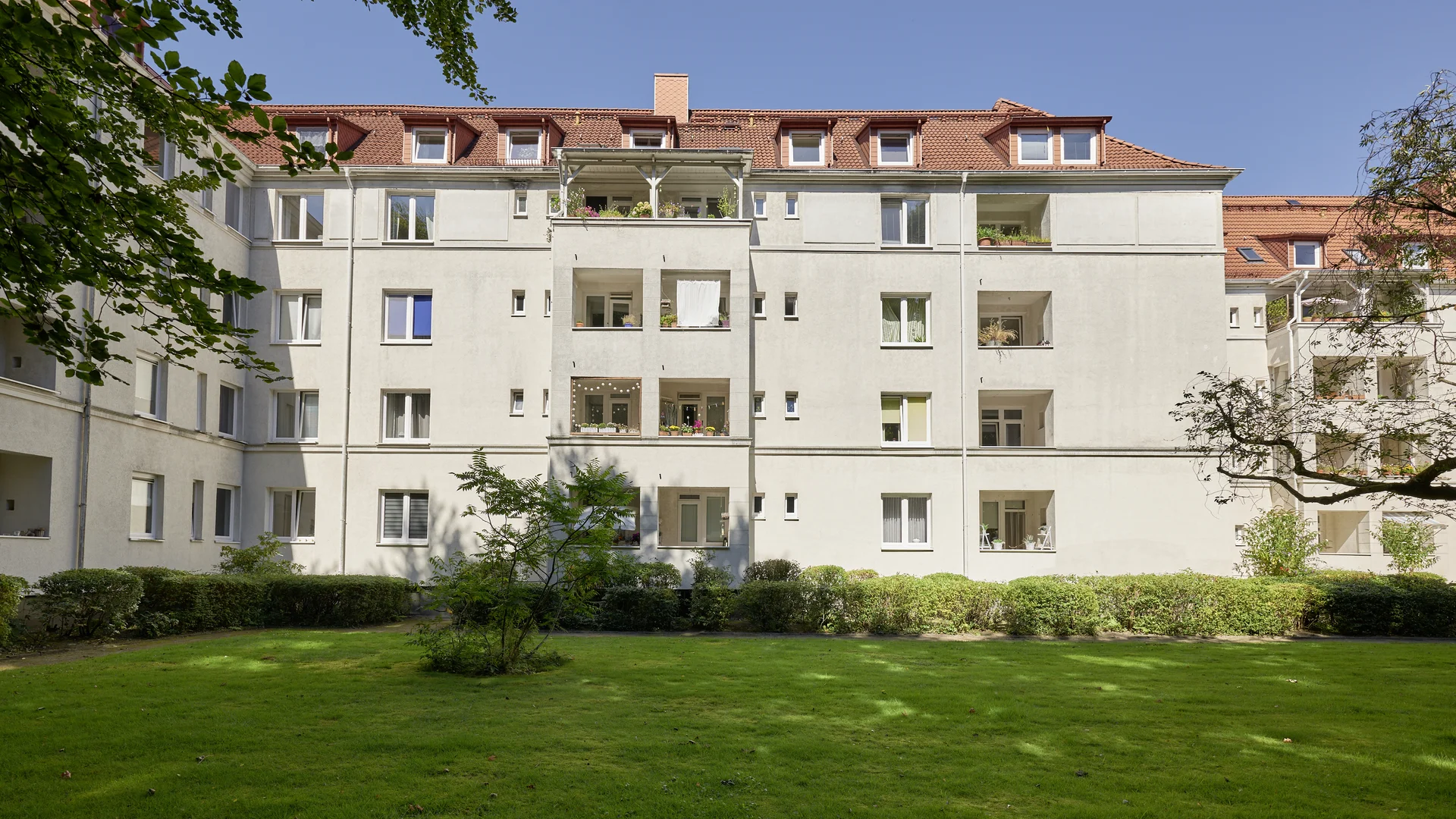 Viergeschossiges Wohnhaus mit heller Fassade, rotem Ziegeldach und Balkonen. Im Vordergrund liegt eine begrünte Innenhoffläche mit Rasen, Hecken und Bäumen.