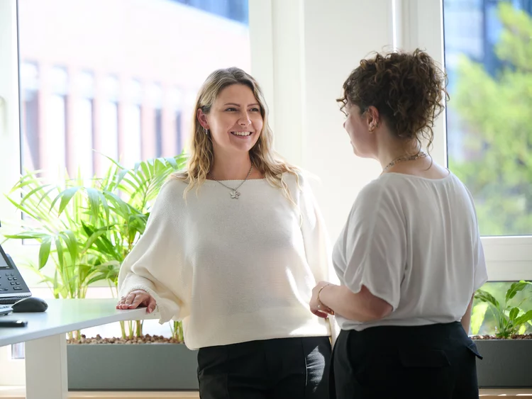 Eine helle, tageslichtdurchflutete Innenaufnahme zeigt zwei lächelnde Frauen, die sich in einem modernen Büro unterhalten. Die eine Frau hat welliges blondes Haar und trägt einen weißen Strickpullover mit weiten Ärmeln. Sie steht an einem Stehschreibtisch und stützt sich mit der Hand ab, während sie die andere Frau ansieht und lacht. Die zweite Frau hat dunkles, lockiges Haar, das sie als Dutt trägt, und trägt eine weiße, locker sitzende Bluse. Sie steht rechts neben der blonden Frau, den Rücken leicht zum Betrachter gewendet, und blickt zu ihr. Auf dem Schreibtisch links stehen ein Computerbildschirm und ein VoIP-Telefon. Hinter den beiden Frauen, vor den großen Fenstern, die den Blick auf ein angrenzendes Gebäude freigeben, befindet sich ein länglicher Blumentopf mit grünen Büropflanzen. Das Licht im Raum ist hell und natürlich.