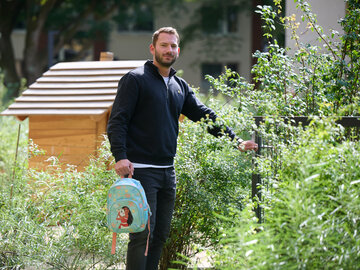 Ein Mann mit dunklem Pullover steht in einem Außenbereich neben einem kleinen Holzhäuschen, hält einen bunt bedruckten Kinderrucksack in der Hand und öffnet ein Gartentor.