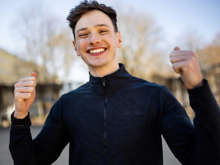 Nahaufnahme eines jungen Mannes im Freien, der breit und glücklich in die Kamera lächelt. Er hat kurze braune Haare, trägt einen dunklen Sport-Oberteil mit Stehkragen und Reißverschluss und ballt beide Fäuste jubelnd auf Schulterhöhe. Der Hintergrund ist unscharf und zeigt Bäume sowie Gebäude im Tageslicht.