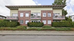 Frontalansicht eines modernen, zweigeschossigen Wohngebäudes mit Klinker- und Putzfassade, Balkonen und dicht bepflanztem Vorgarten.