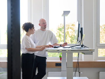 Eine Frau und ein Mann stehen gemeinsam in einem hellen Büro an einem höhenverstellbaren Schreibtisch und blicken auf einen Computerbildschirm.