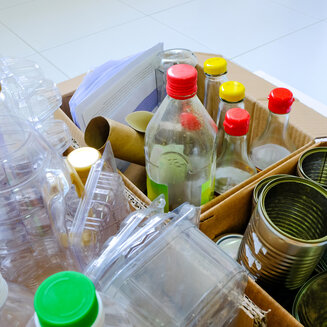 Nahaufnahme von für das Recycling sortiertem Abfall in Pappkartons. Im Vordergrund liegen transparente Plastikflaschen und Kunststoffverpackungen, daneben Glasflaschen mit bunten Deckeln sowie leere Konservendosen aus Metall.