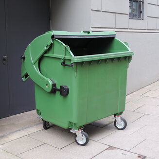 Ein großer grüner Müllgroßbehälter aus Kunststoff steht auf einem gepflasterten Gehweg direkt vor einer grauen Hauswand. Der Schiebedeckel des Containers ist einen Spalt weit nach hinten geöffnet.