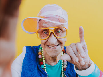 Eine ältere, fröhlich lächelnde Frau macht ein Selfie vor gelbem Hintergrund. Sie trägt eine orangefarbene Sonnenblende, eine auffällige gelbe Brille, buntes Zubehör und macht eine lässige Handgeste.