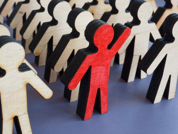 Viele kleine Holzfiguren stehen nebeneinander, während eine einzelne, rot eingefärbte Figur mit erhobenem Arm aus der Masse hervorsticht.