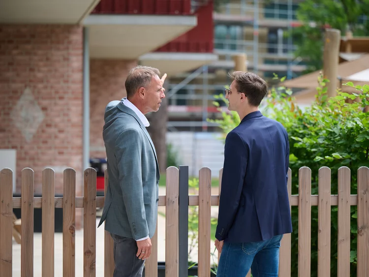 Rückenansicht von Stefan Henze (links) und Eric Yeung-Schüürmann (rechts), die an einem Holzzaun stehen. Stefan Henze zeigt mit ausgestrecktem Arm auf ein eingerüstetes Bauprojekt im unscharfen Hintergrund.