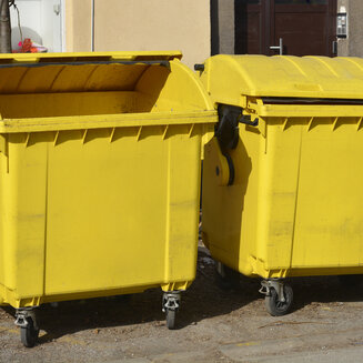Zwei große gelbe Plastikmüllcontainer auf Rädern stehen nebeneinander auf einem Gehweg vor einer Häuserwand. Der linke Container hat einen weit geöffneten Runddeckel, der rechte ist geschlossen.