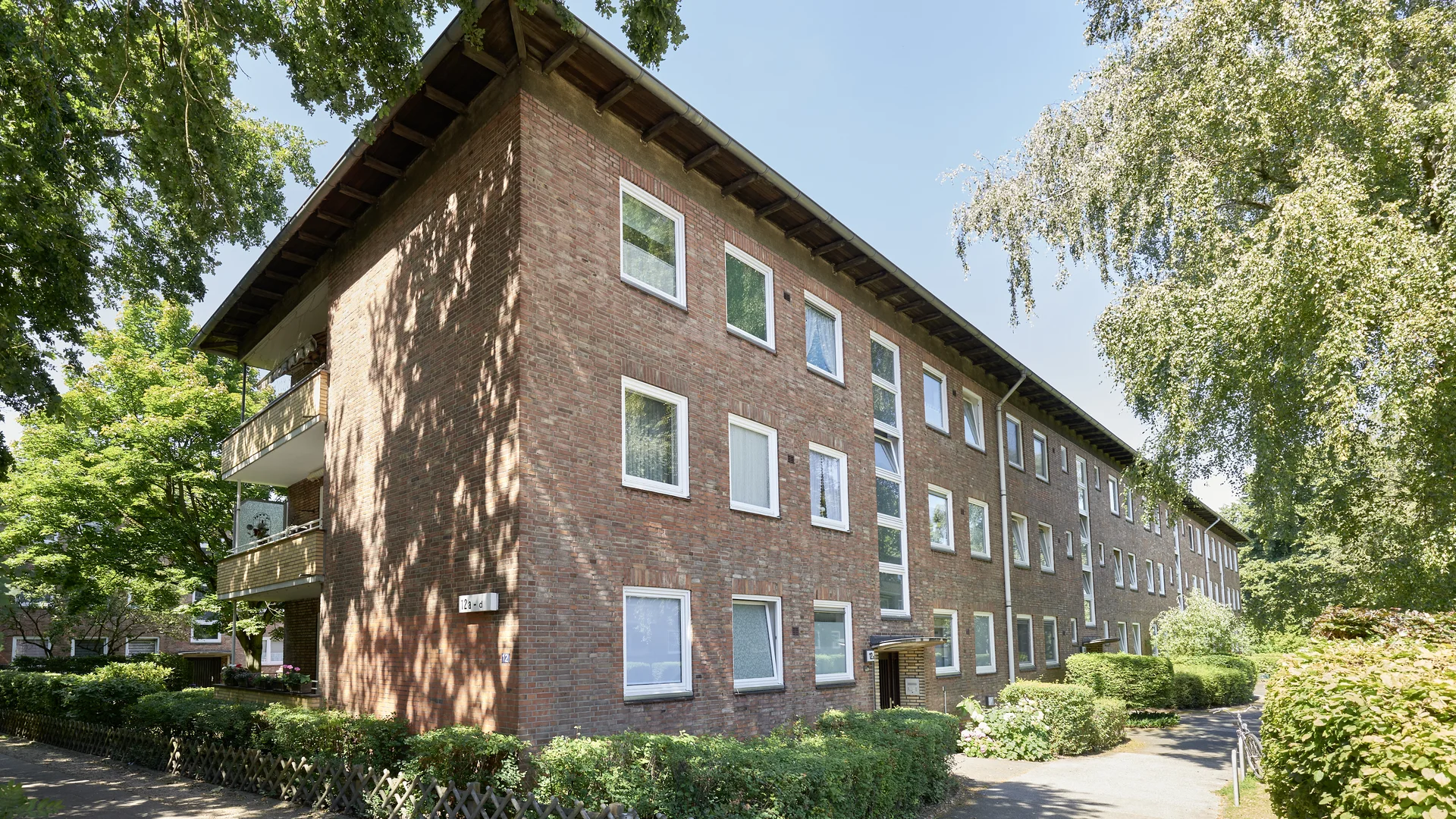 Dreigeschossiges Wohnhaus mit roter Backsteinfassade und Balkonen, umgeben von Bäumen, Hecken und begrünten Wegen.
