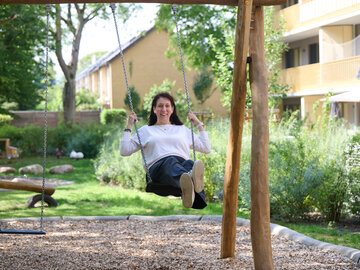 Eine lächelnde Frau mit dunklen Haaren schaukelt auf einem Kinderspielplatz im Grünen vor einer Wohnanlage.