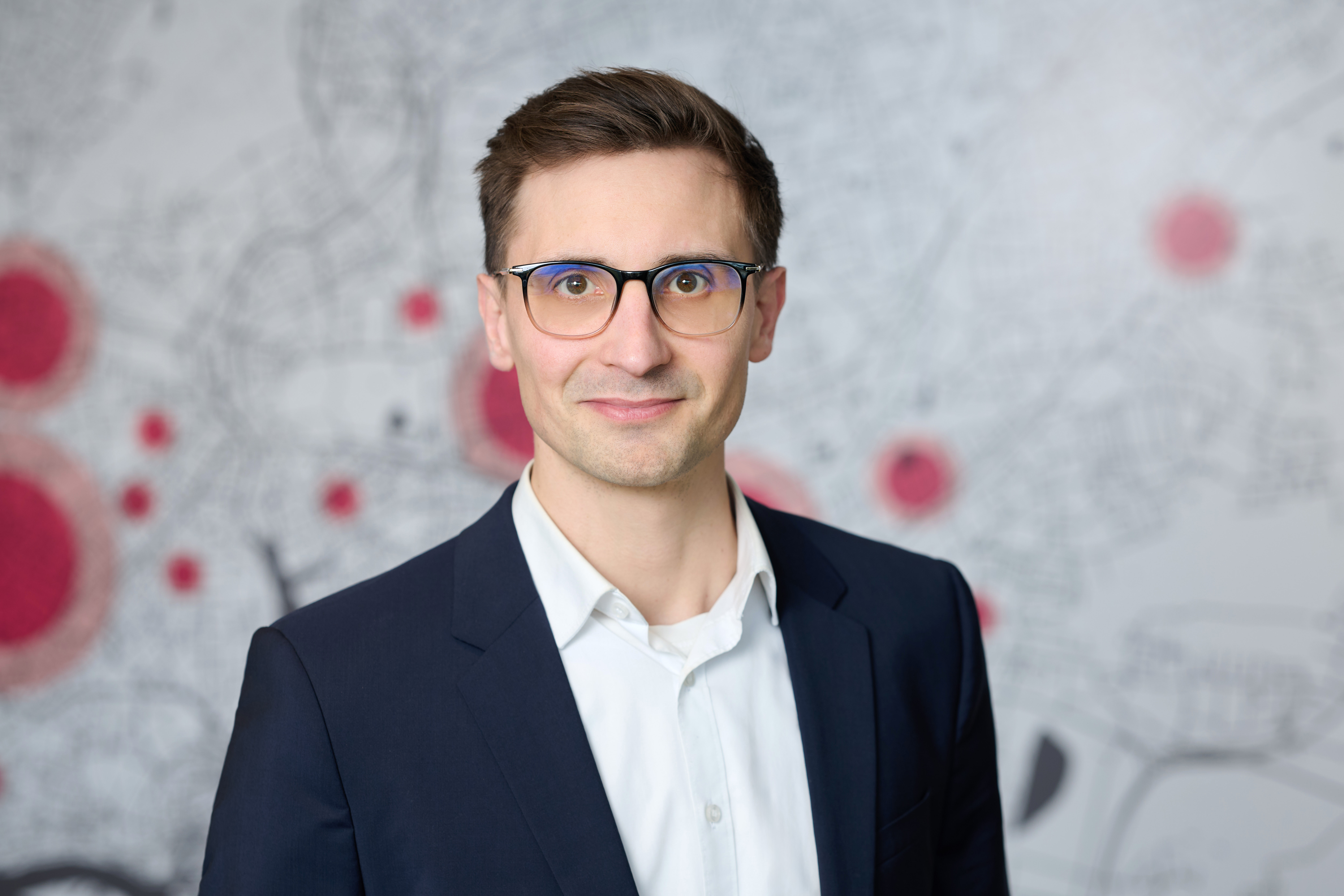 Ein junger Mann mit kurzen braunen Haaren, Eric Yeung-Schüürmann, blickt lächelnd in die Kamera. Er trägt eine Brille mit dunklem Rahmen, ein weißes Hemd und ein dunkelblaues Sakko. Im unscharfen Hintergrund ist ein schematischer Stadtplan in Grau mit mehreren roten Markierungspunkten zu sehen.